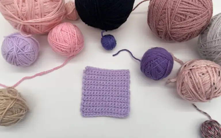 Gehäkeltes lilafarbenes Quadrat, umgeben von Wollknäueln in verschiedenen Farben.