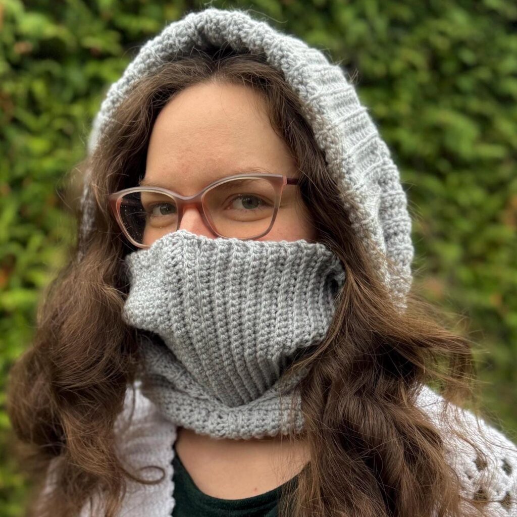 Frau mit Brille trägt eine graue gestrickte Kapuze und Schal, umgeben von einem grünen Hintergrund