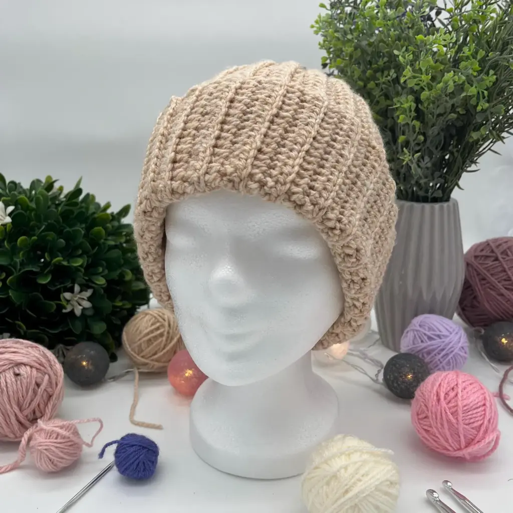 Eine Strickmütze in Beige auf einem weißen Schaumstoffkopf, umgeben von Garnknäueln und Pflanzen