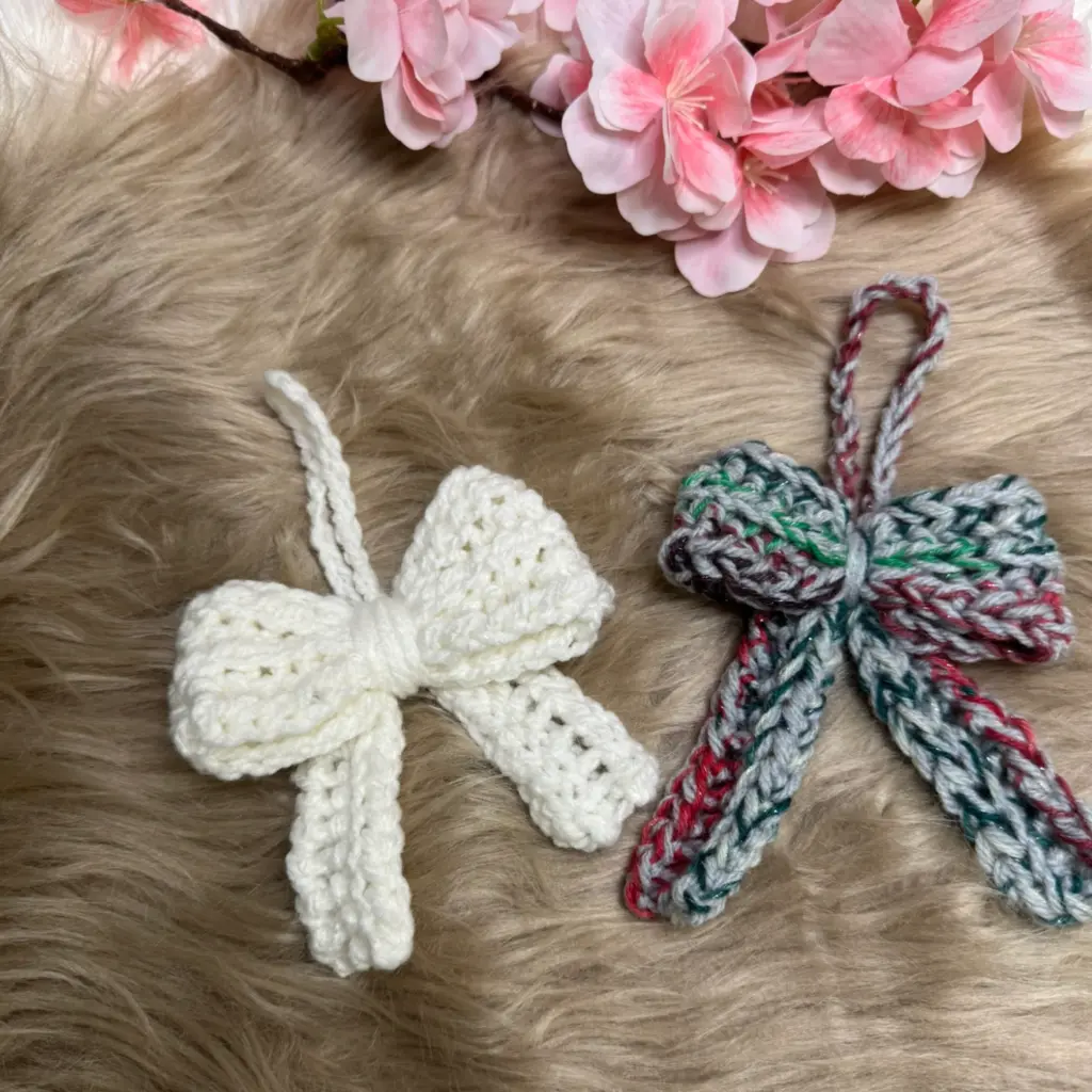 Zwei gehäkelte Schleifen, eine in Weiß und die andere mehrfarbig, liegen auf einem flauschigen Untergrund, daneben rosa Blüten