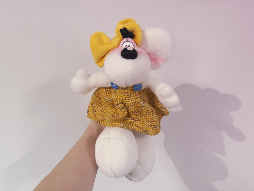 Plüschfigur einer weißen Maus mit gelber Schleife und gestricktem, gelben Kleid, gehalten von einer Hand